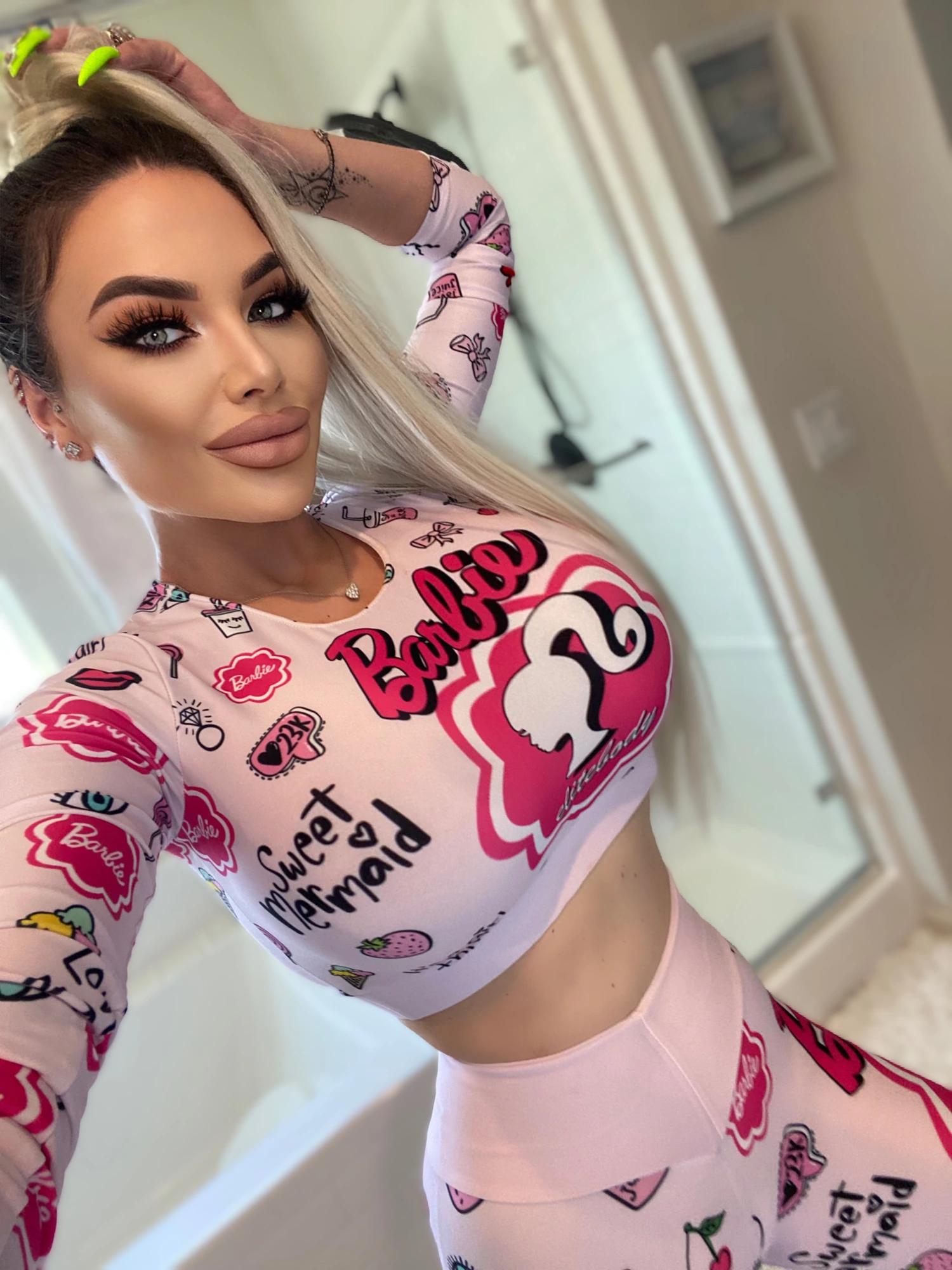 A selfie of Sasha in a pink Barbie-print crop top and leggings, one arm raised behind her head and her long blonde hair over one shoulder.