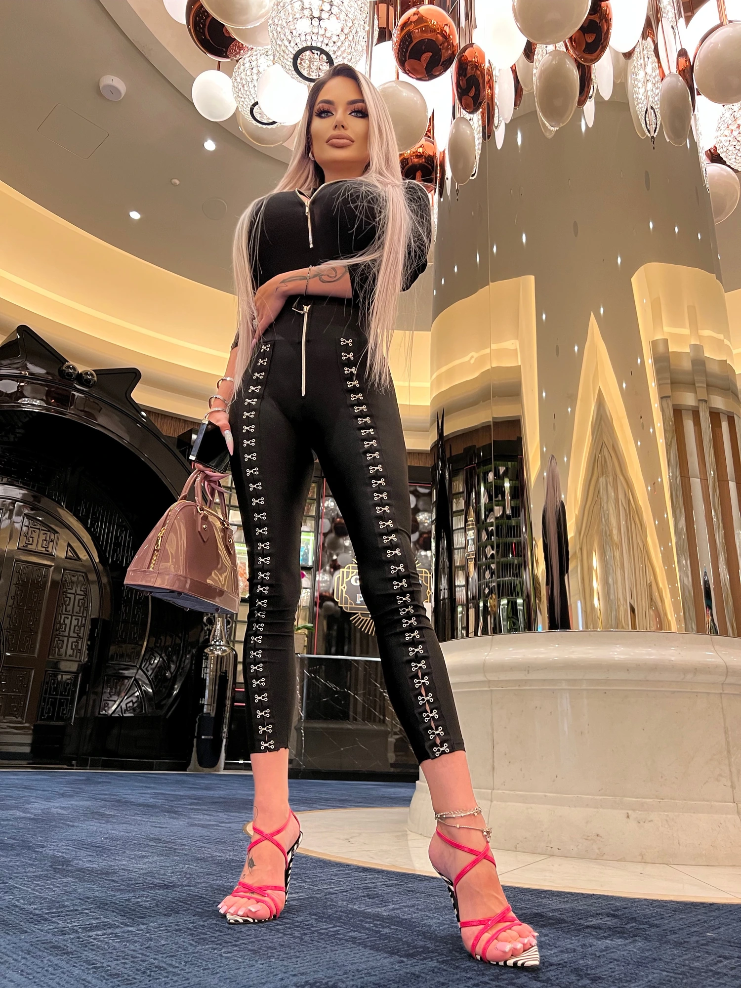 Sasha in a black zip-front jumpsuit trimmed with silver hooks and pink strappy heels, in a grand casino lobby under a chandelier of glass globes.
