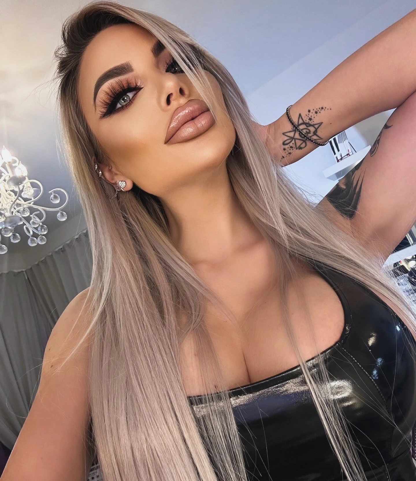 Sasha in a black patent top, one arm raised to show a star tattoo on her wrist, with a crystal chandelier behind her.