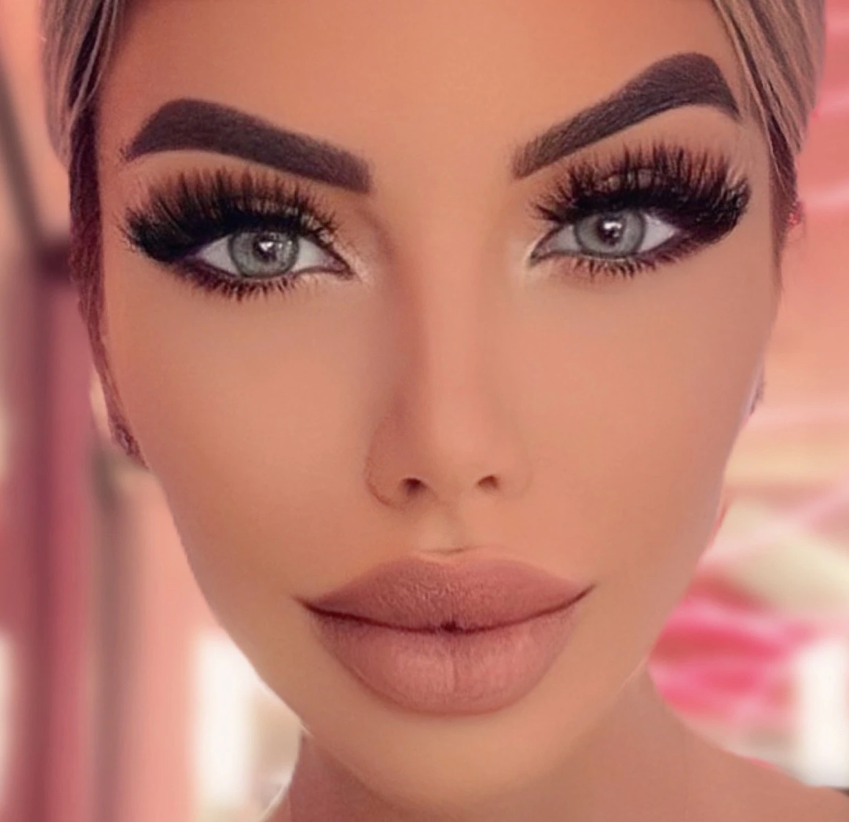 Sasha looking straight into the camera against a soft pink blur: bold brows, dramatic lashes and a peachy nude lip.