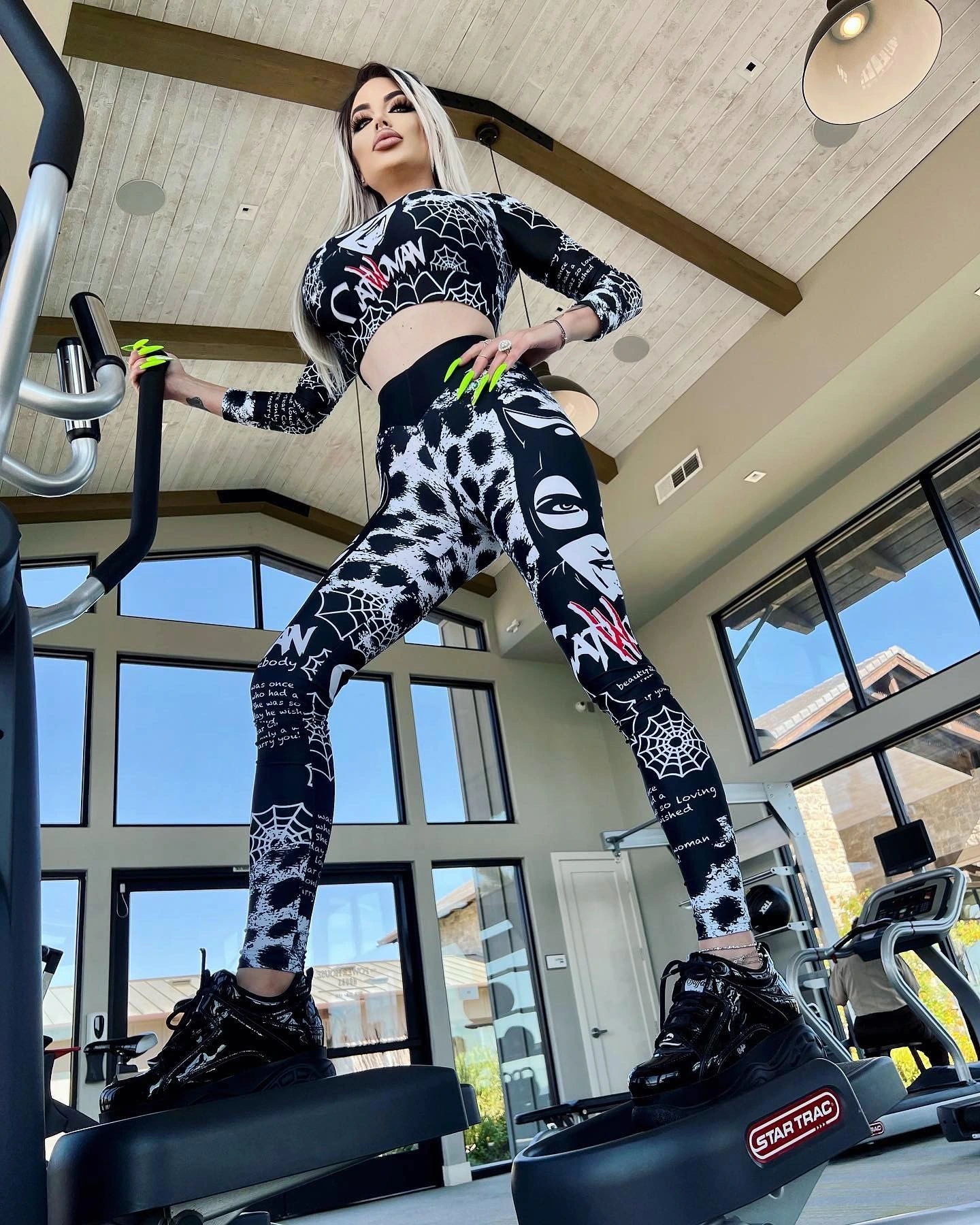 Sasha seen from below on a stair climber under a wood-beamed ceiling, in a black and white printed crop top and leggings and chunky black sneakers.