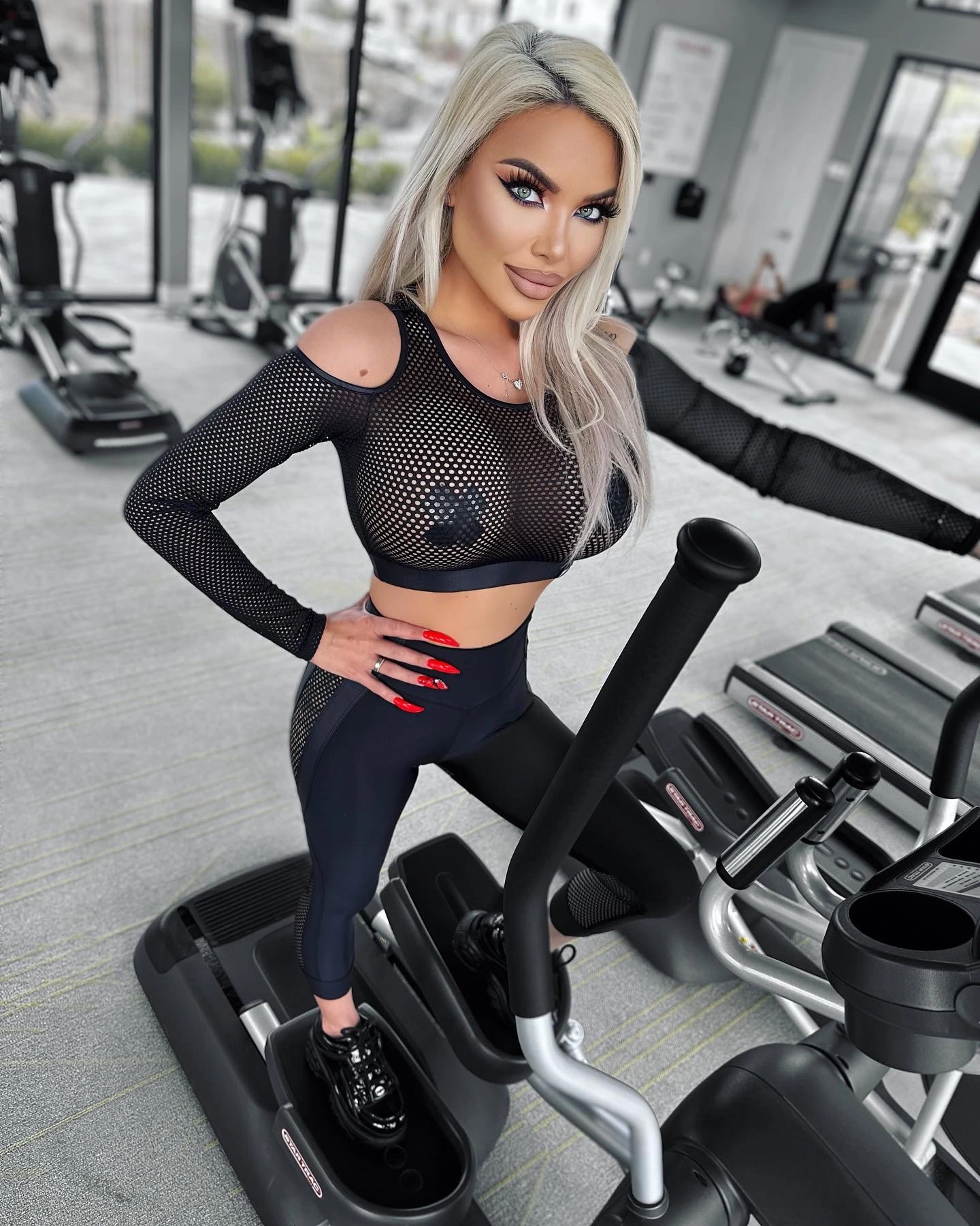 Sasha on an elliptical in a bright gym, one hand on her hip, in a black mesh crop top with open shoulders, black leggings and black patent platform sneakers.
