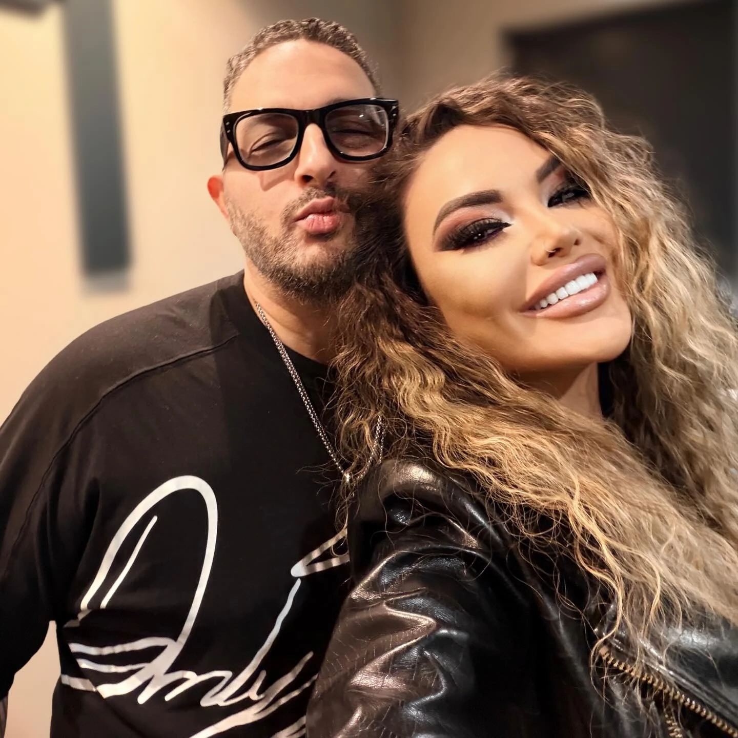 Sasha and Adam cheek to cheek, smiling, him in glasses and a black tee, her in a leather jacket.