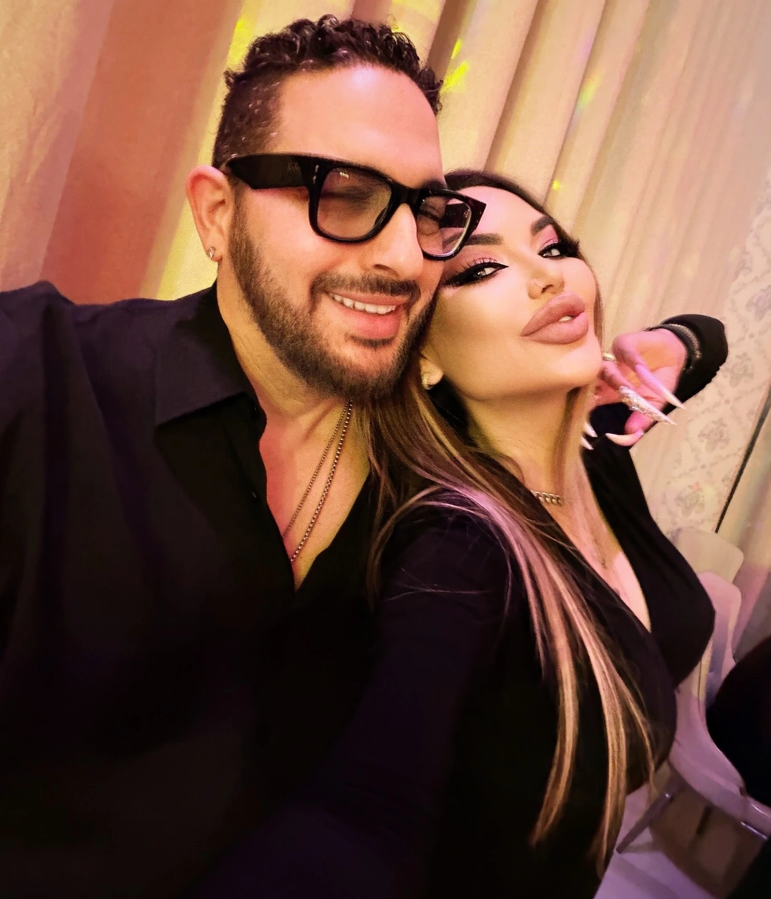 Sasha and Adam on a night out, both in black, close together under warm pink light.