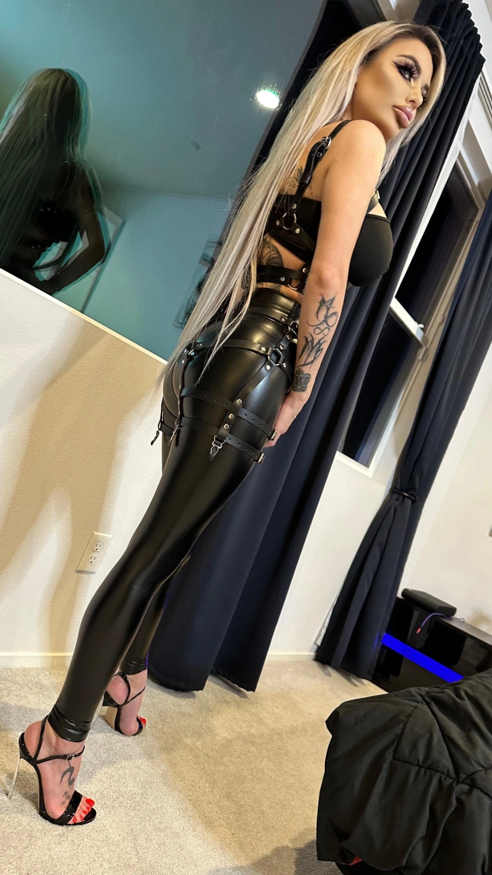 Sasha from behind in high-shine black leggings with buckled straps and strappy heels, looking back over her shoulder.