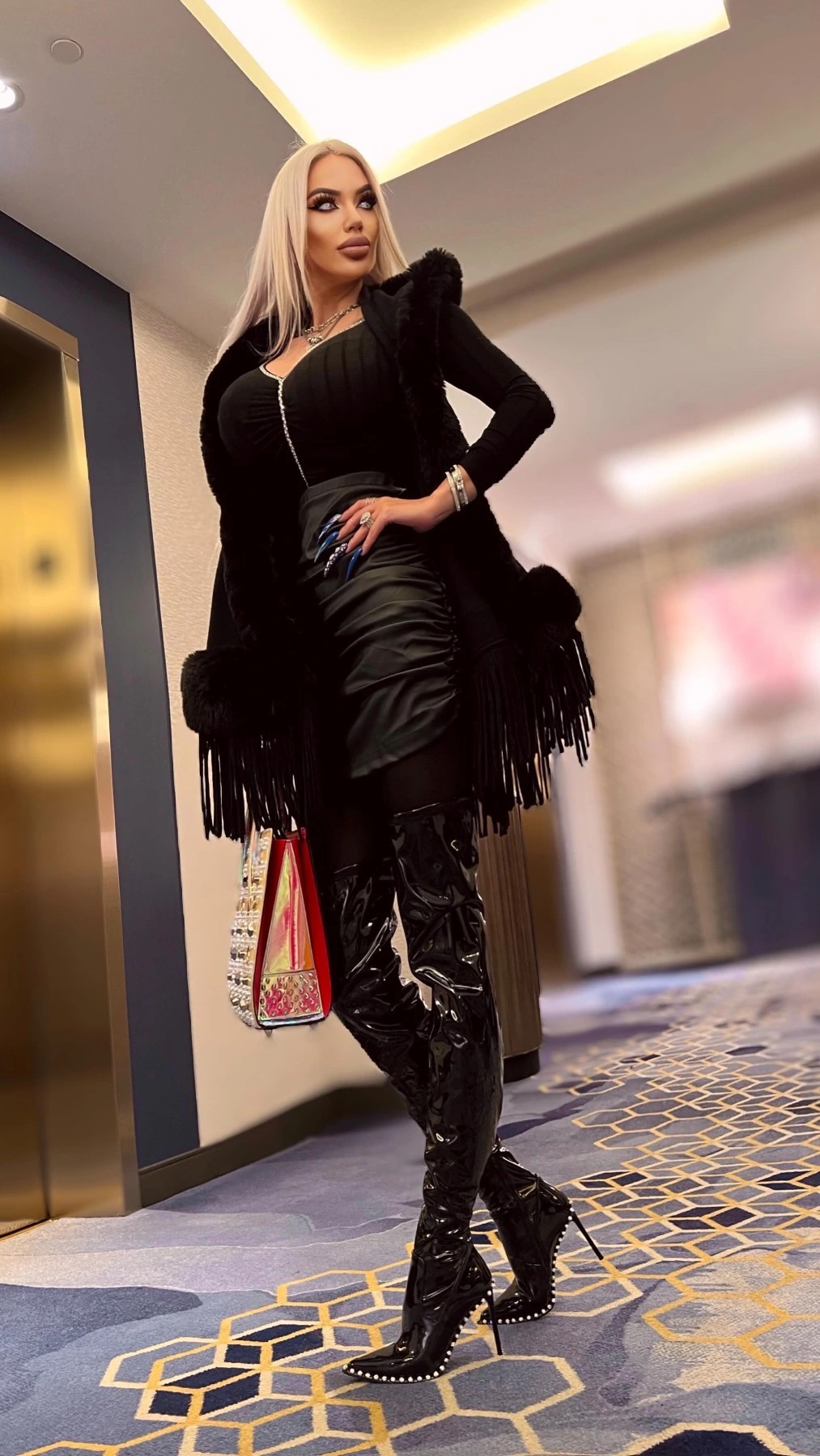 Sasha head to toe in black in a hotel corridor: a fringed shawl over a ribbed top and leather mini, patent thigh-high boots with studded stiletto heels.