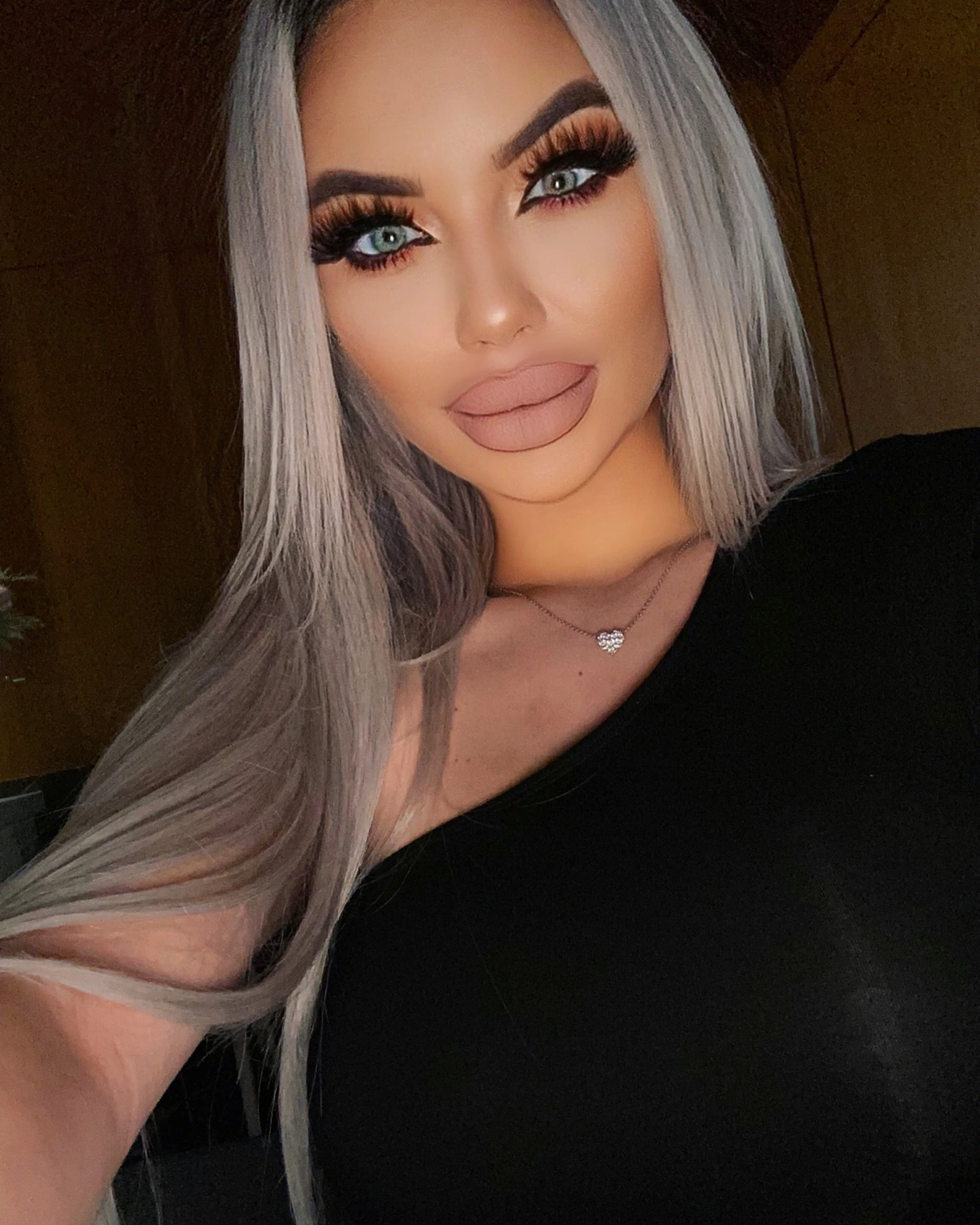 A warm close-up of Sasha in a black one-shoulder top and a small diamond heart pendant, her long silver-blonde hair falling past her shoulders.