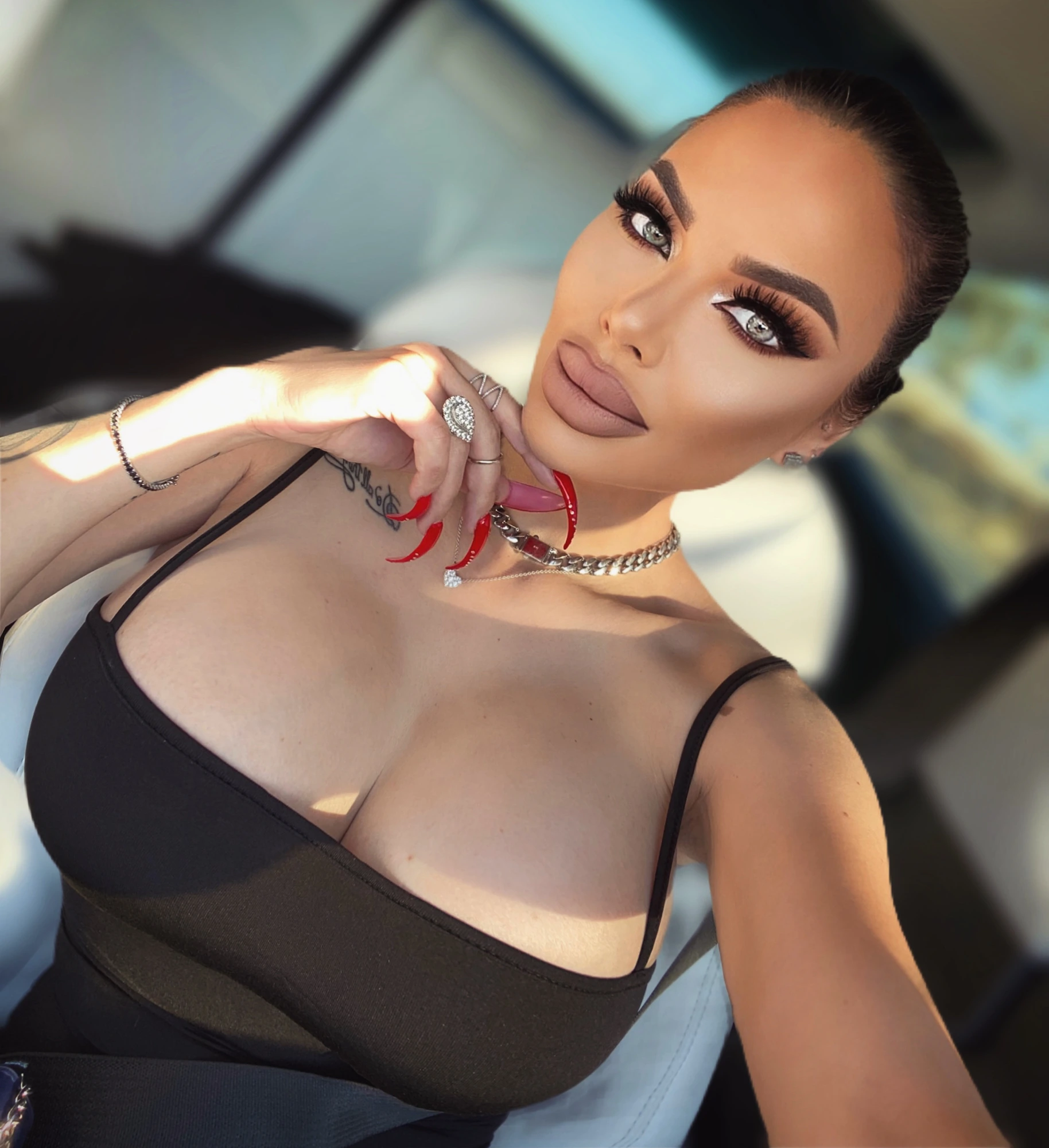 Sasha with her hair pulled back sleek, long red stiletto nails at her chin, a diamond ring and a chain necklace, in a black top.