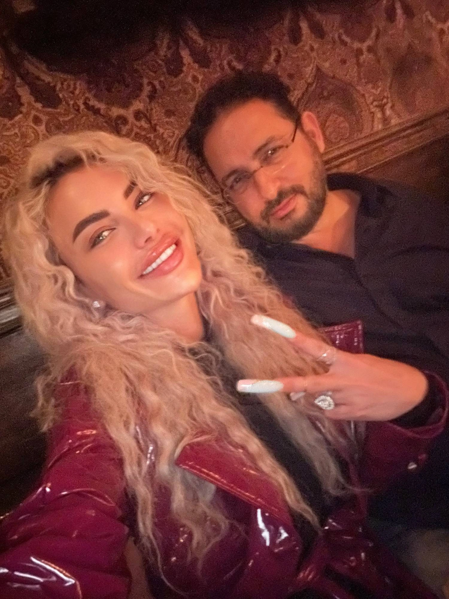 Sasha and Adam in a velvet booth on a night out, her long curly blonde hair over a burgundy patent coat, smiling and making a peace sign with long white nails.