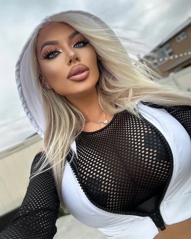 A close selfie of Sasha outdoors, a white hood up over her long blonde hair and a black mesh top beneath.